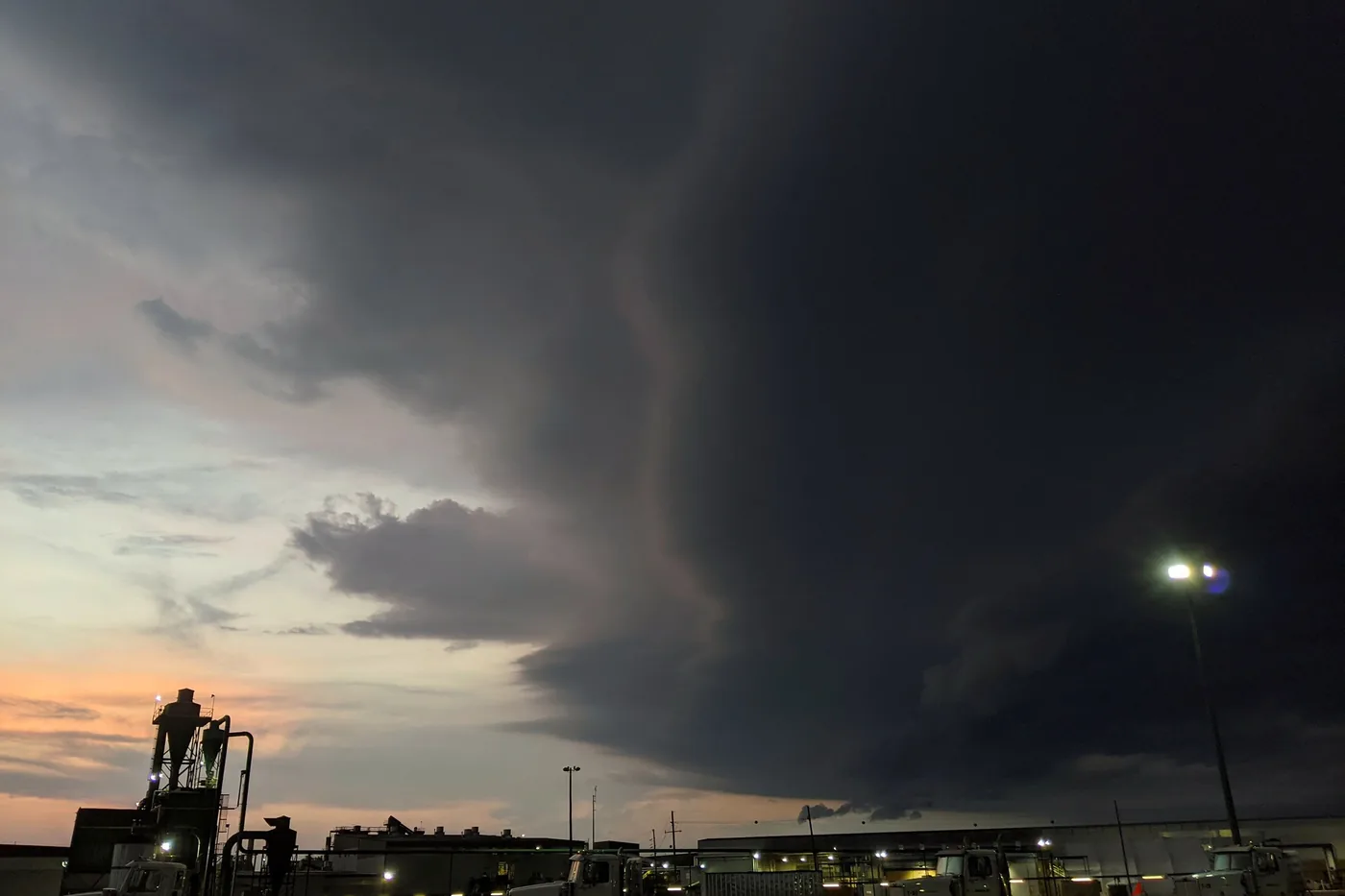 Dark thunderstorm cloud towering over a lit industrial yard at dusk in central Louisiana