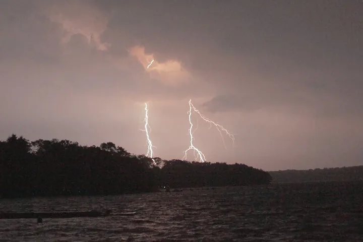 Lightning bolts striking from a storm over the water along a wooded Rhode Island shoreline at dusk